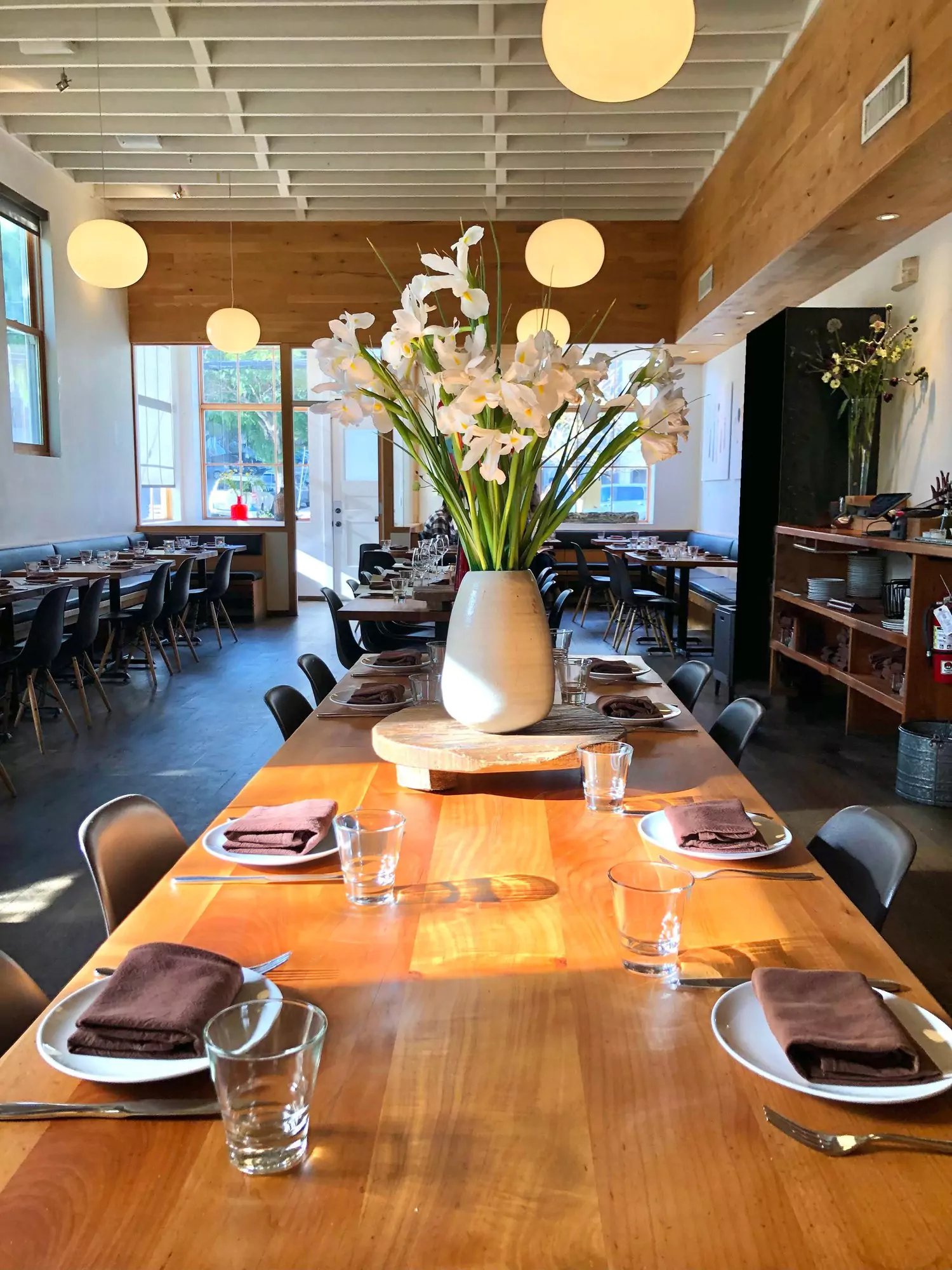 Interior contemporary casual dining room with long communal wood dining tables, a large white vase filled with white flowers, place settings including brown cloth napkins, and black mid century modern chairs