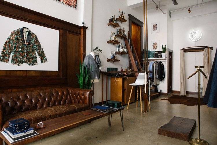 Interior of Tailors’ Keep Shop in San Francisco with Tufted Brown Leather Sofa, Long Modern Wood Coffee Table, Patterned Suit Jacket Sketch above Sofa, and other Bespoke Men’s Clothing and Accessory Items in the Background