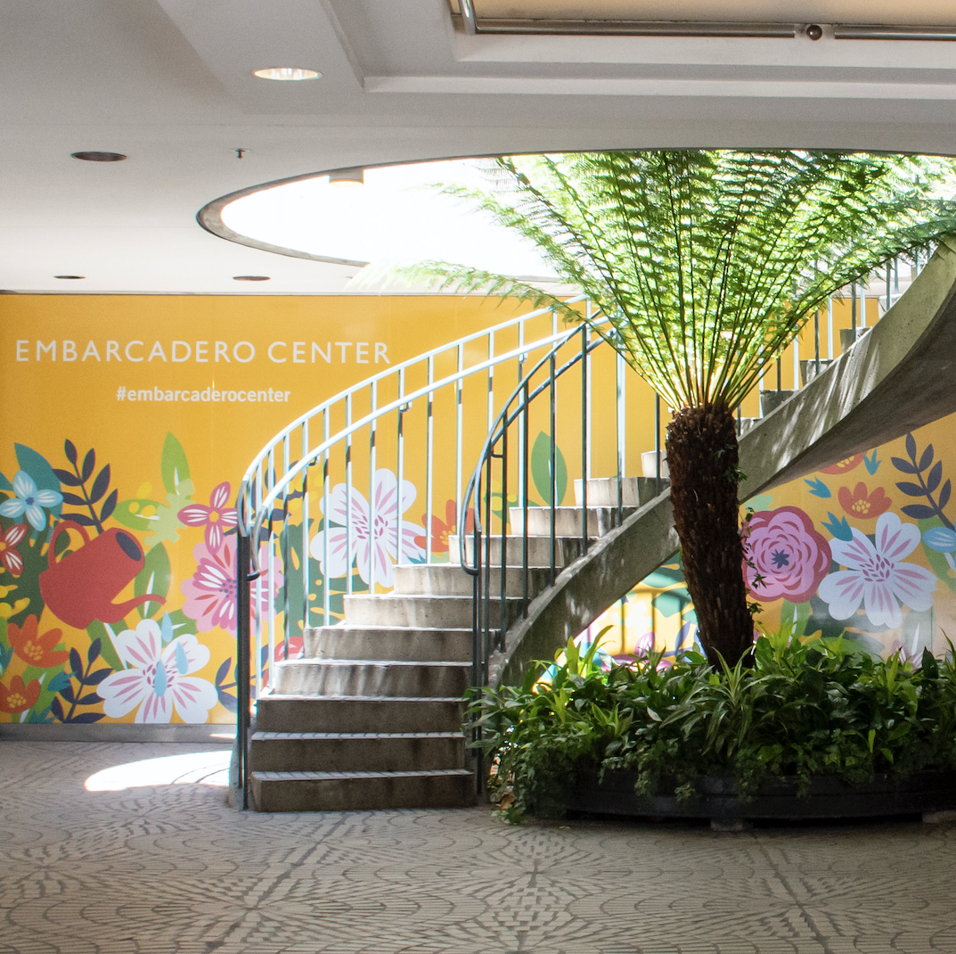 Wall Art with Yellow Background and Flowers with Watering Can that Says Embarcadero, Behind a Curving Outdoor Staircase and Palm Tree